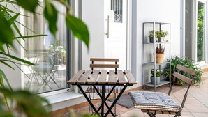 Un patio con una mesa y sillas de madera, una puerta de cristal, un estante de metal con plantas y una ventana.