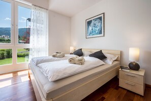 Un lit double avec du linge de lit blanc, une table de chevet avec une lampe, un tableau encadré au mur et une vue sur la verdure par la fenêtre.