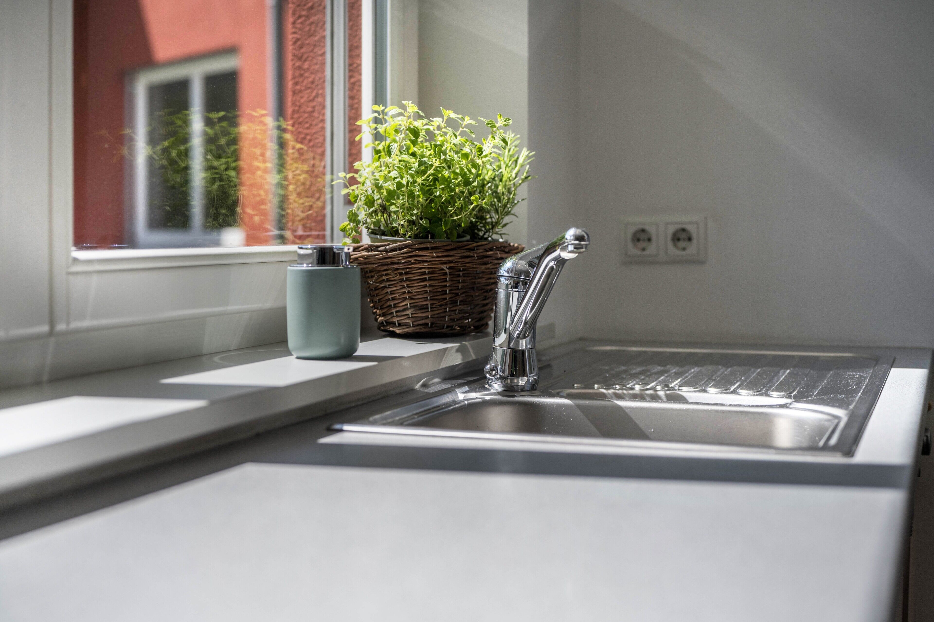 Een keuken met een roestvrijstalen spoelbak, een rieten mand met groene planten en een raam met uitzicht op een gebouw.