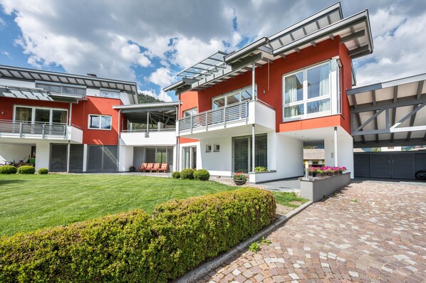 Une maison moderne à deux étages, dotée d’une façade rouge, de grandes fenêtres et d’une pelouse bien entretenue.