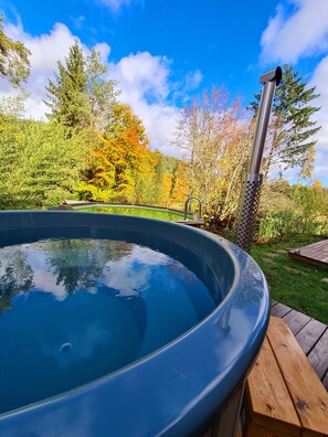 Un bain à remous entouré d’une terrasse en bois, avec une structure métallique sur la droite, le tout se détachant sur un fond d’arbres et d’un ciel dégagé.