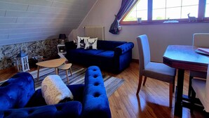 Una habitación estilo loft con techo inclinado, un sofá azul, una mesa de comedor y vistas a través de un ventanal grande.