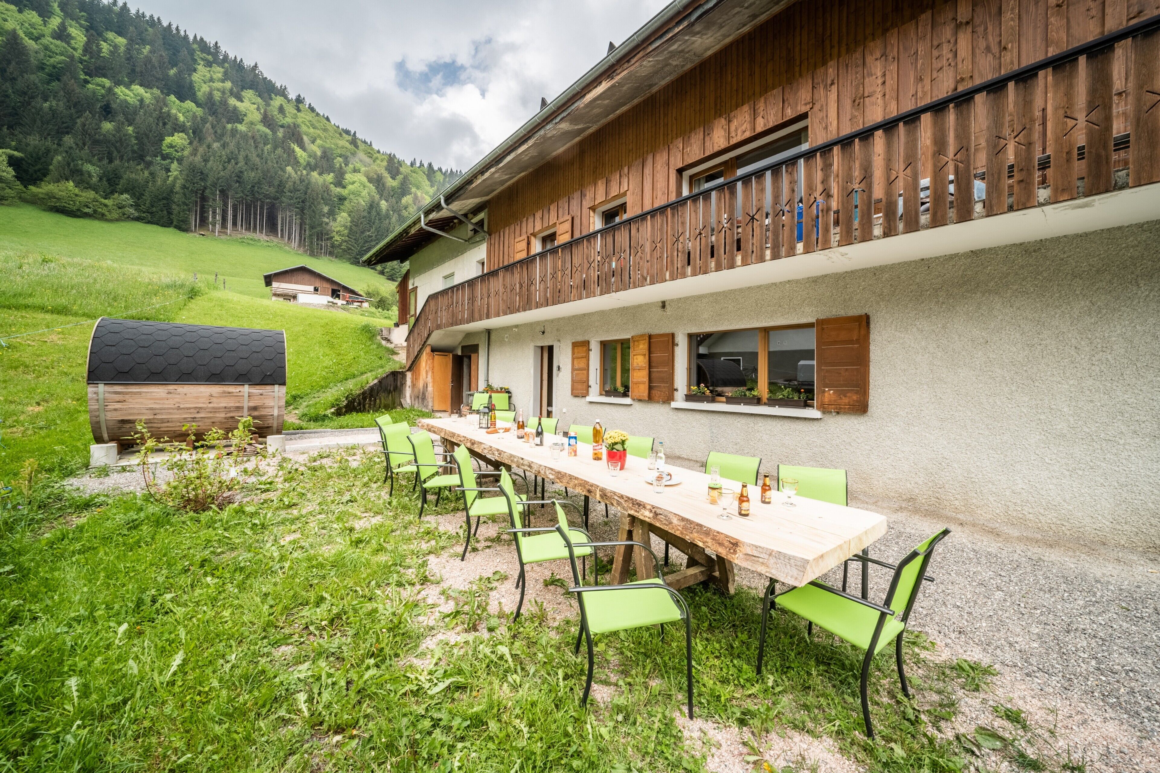 Et langt spisebord med stole, placeret udendørs med udsigt til et bjerg og en træbygning.