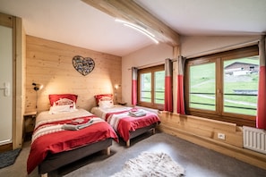 Dormitorio de cabaña de madera con dos camas, una decoración de corazón en la pared y vistas a un paisaje verde a través de un ventanal grande.