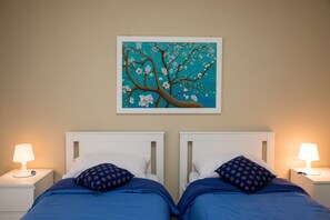 A room with two beds, each with a white headboard, a blue bedspread, and a patterned pillow. There is a wall-mounted painting of a tree with white blossoms.
