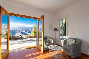 Une pièce dotée d’une grande porte-fenêtre coulissante donnant sur une terrasse avec une table et des chaises, deux fauteuils gris, une petite table ronde blanche et une vue sur les montagnes.