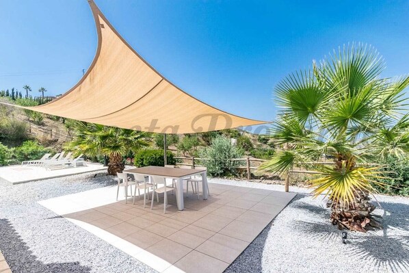 Une terrasse extérieure avec une table et des chaises, une grande voile d’ombrage et un palmier.