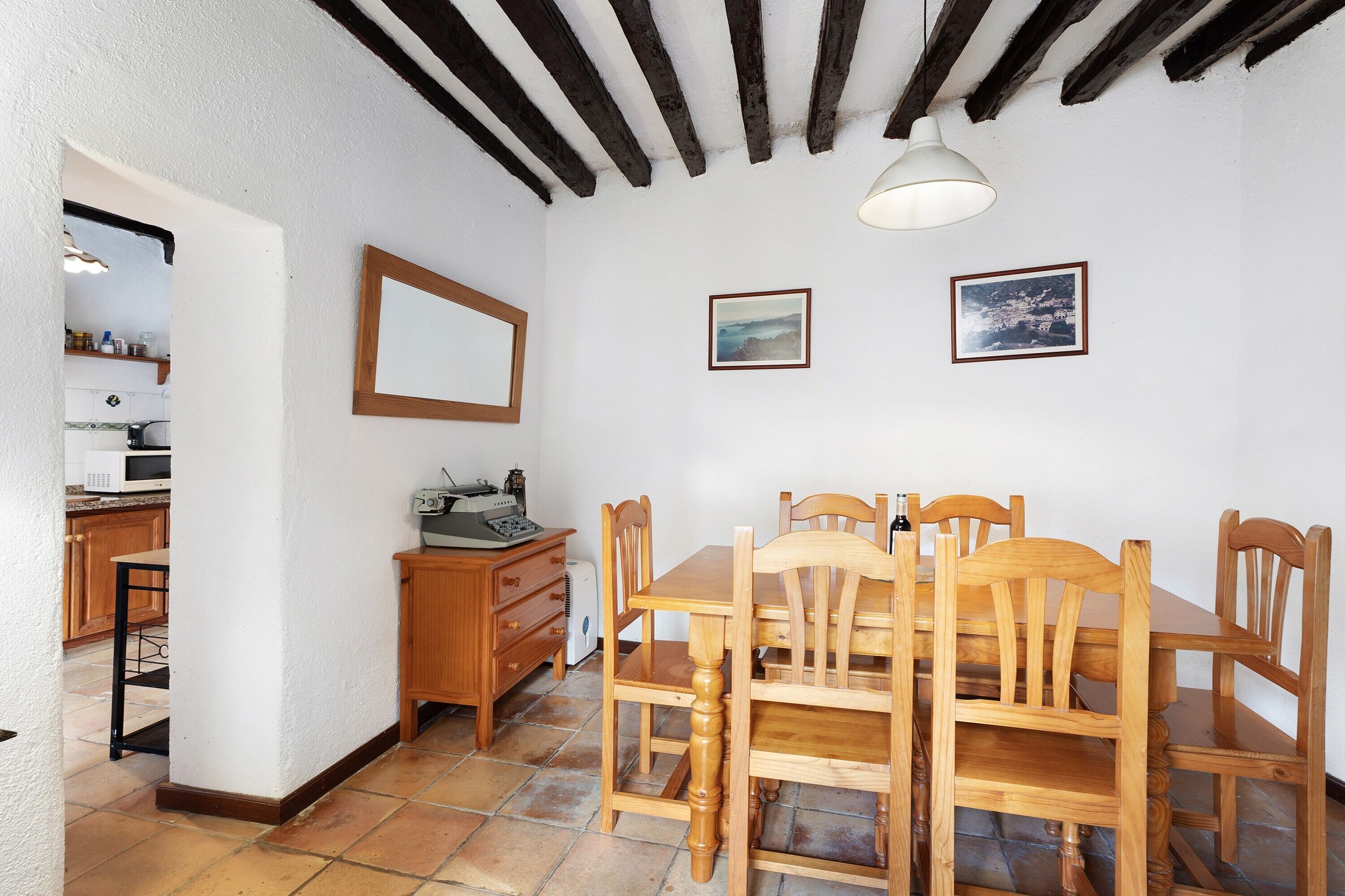 Un coin repas avec une table et des chaises en bois, un mur blanc orné de deux tableaux encadrés, et une cuisine visible à travers une porte ouverte.