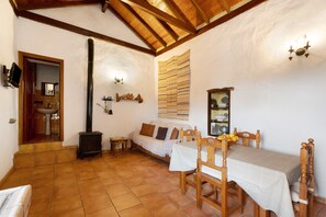A cozy room with a wooden ceiling, a sofa, a dining table with chairs, a fireplace, and a wall-mounted shelf with dishes.