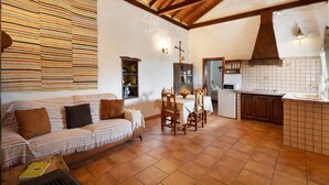 A cozy living room with a sofa, dining table, and chairs, a kitchen area with a tiled backsplash, and wooden beams on the ceiling.