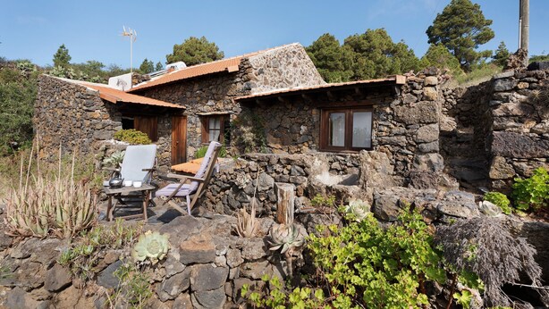 A stone house with a sloped roof, a small porch with a chair, and a wooden door.
