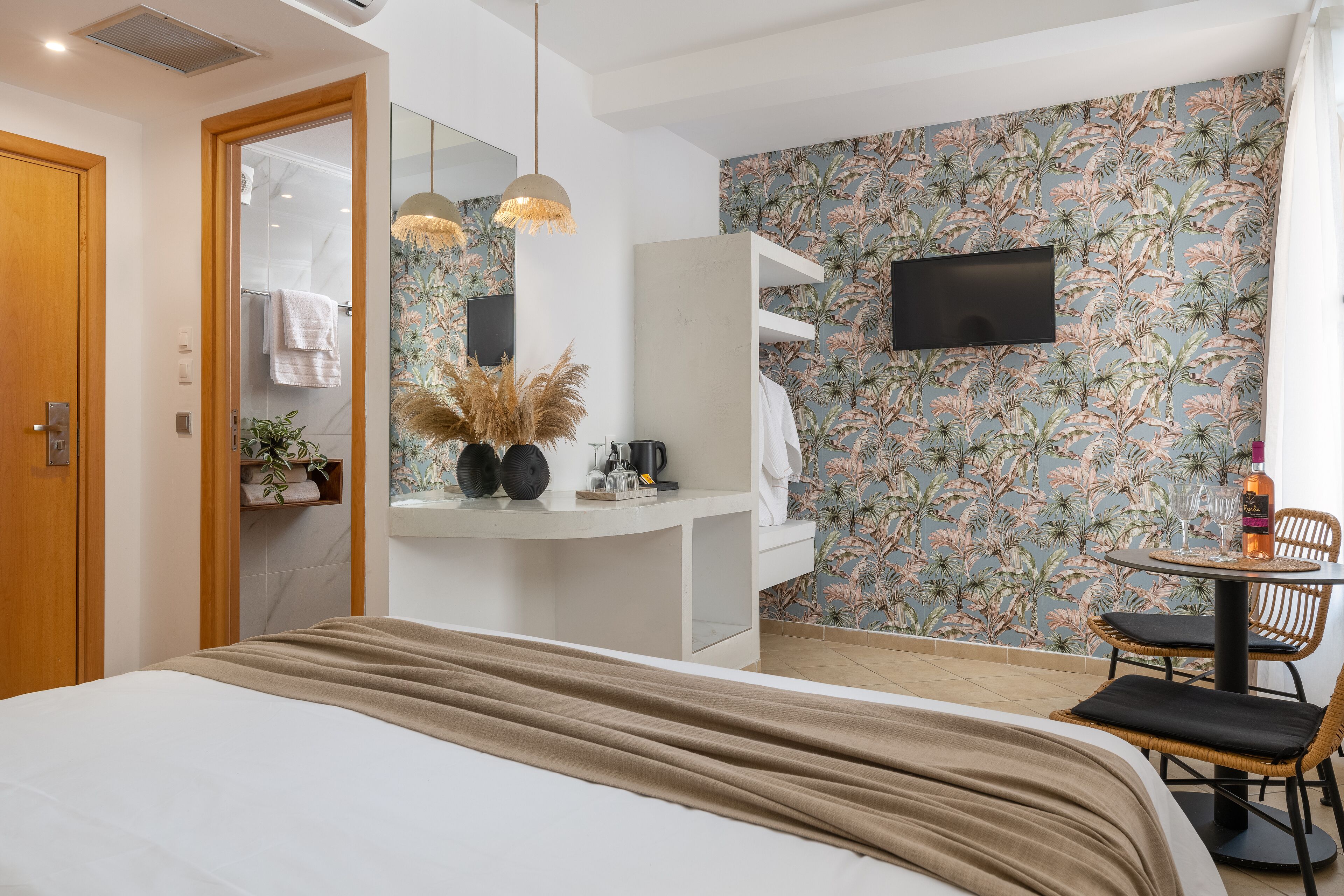 A modern hotel room with a bed, a small table with chairs, a TV, a bathroom with a shower, and a floral wallpaper.