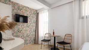 Ein Hotelzimmer mit einer weißen, gebogenen Rezeption, zwei Korbstühlen, einem kleinen runden Tisch mit Flasche und Gläsern, einem Flachbildfernseher an einer Wand mit Blumenmuster und einem Bett mit weißer Bettwäsche.