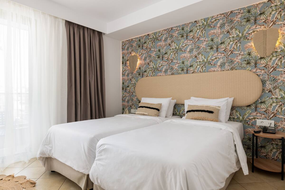 Ein Hotelzimmer mit zwei Betten, floralem Wandpapier, einem großen Spiegel und einem Nachttisch mit Telefon.