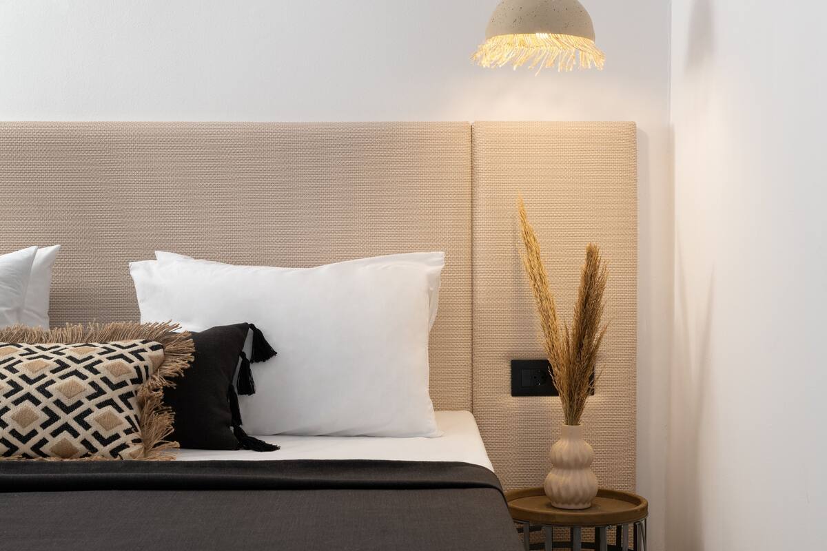 Ein ordentlich gemachtes Bett mit weißen und gemusterten Kissen, ein Nachttisch mit einer Vase mit Trockenpflanzen und eine strukturierte Wand.