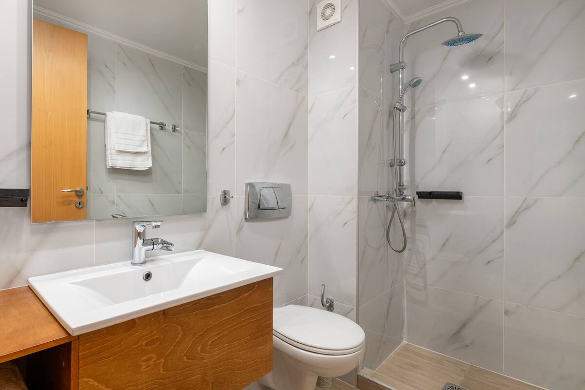 Ein Badezimmer mit weißem Waschbecken, Holz-Badmöbeln, einer Toilette und einer Duschkabine mit Handbrause.