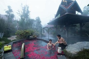 Une source thermale agrémentée de pétales de rose, une structure en pierre et un bâtiment doté d’un toit en pente.