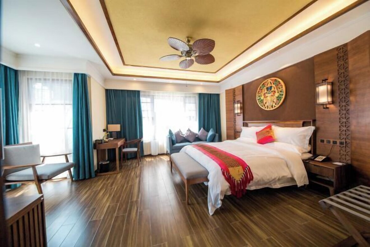Une chambre d’hôtel avec un grand lit, un bureau et une chaise. On y trouve un ventilateur de plafond et un luminaire fixé au mur.