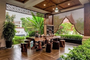 Una mesa de comedor de madera con taburetes, rodeada de plantas en macetas y vistas al verdor exterior.