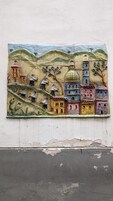 A colorful mosaic mural on a wall depicting a village scene with buildings, trees, and a church.