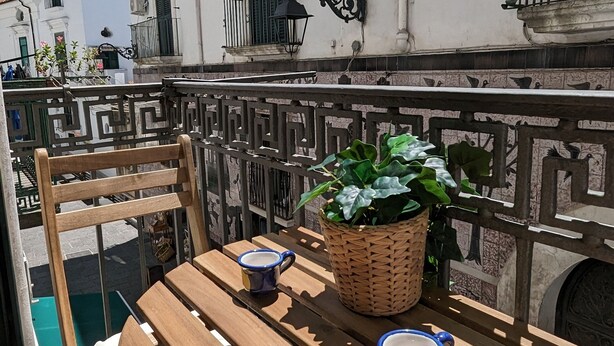 Una mesa de madera con dos tazas azules, una cesta de mimbre y una planta en maceta en un balcón con baranda de hierro forjado.