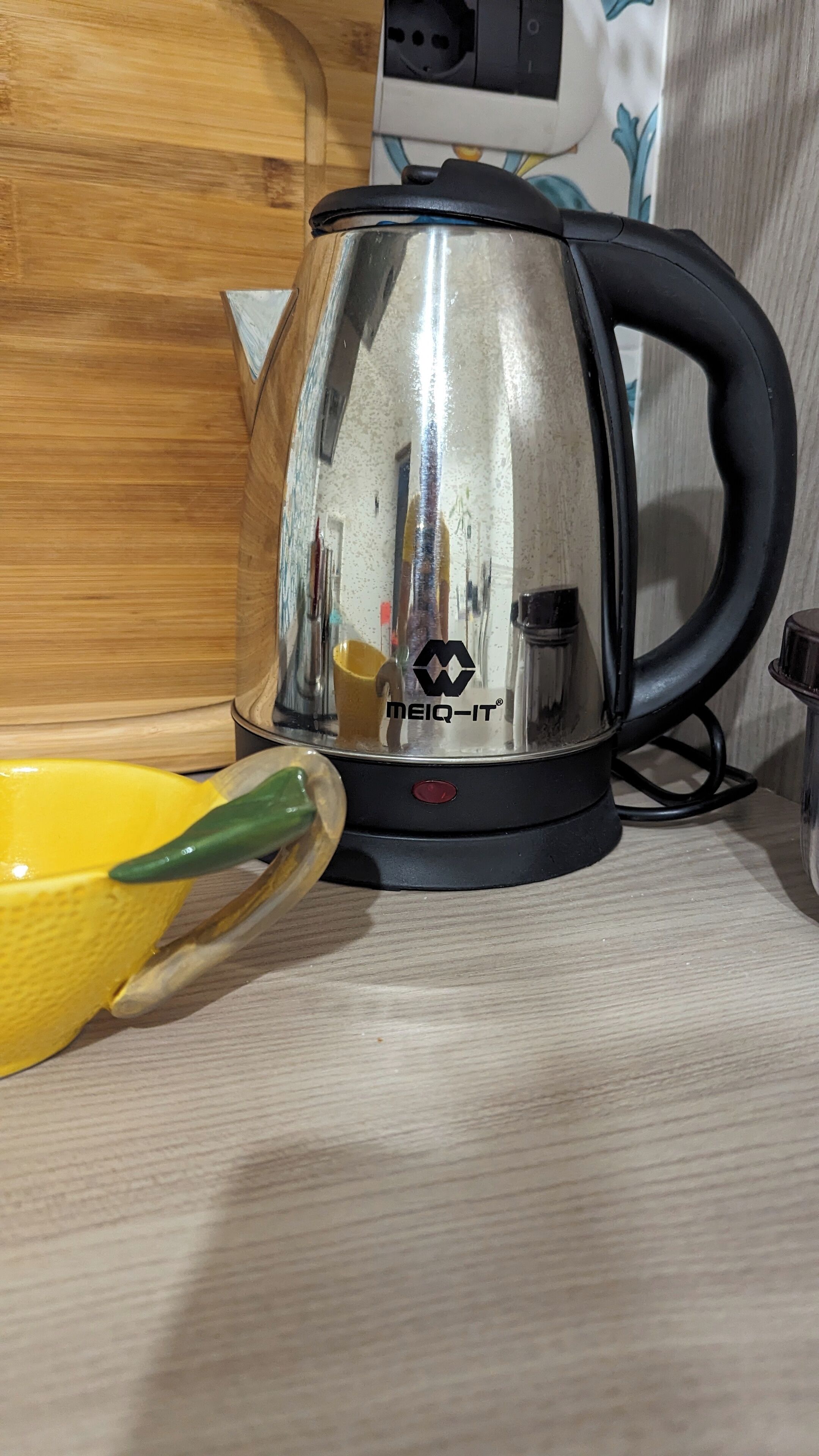 A transparent electric kettle with a black handle and a red button, placed on a wooden surface.