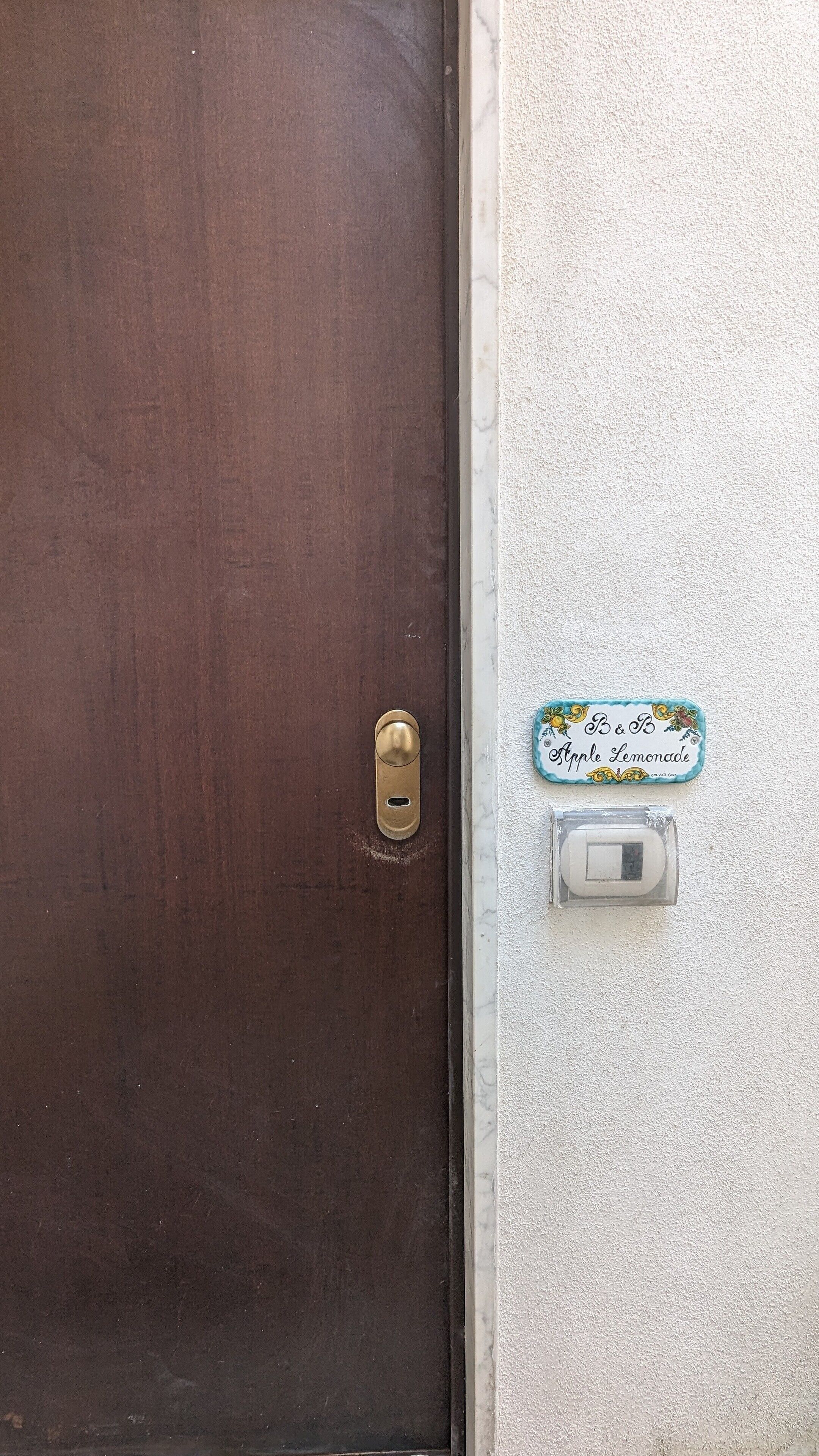Una puerta marrón con un tirador de latón y una cerradura, una pared blanca con textura, y un pequeño cartel que dice "Apple Lemonade".