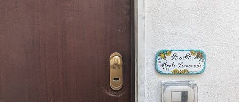 Una puerta marrón con un tirador de latón y una cerradura, una pared blanca con textura, y un pequeño cartel que dice "Apple Lemonade".