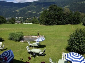 An outdoor seating area with tables and chairs, a small pond, and a scenic view of mountains and a village in the background. - Appartement Gerlitzen - Pension Strauß (Rappitsch)