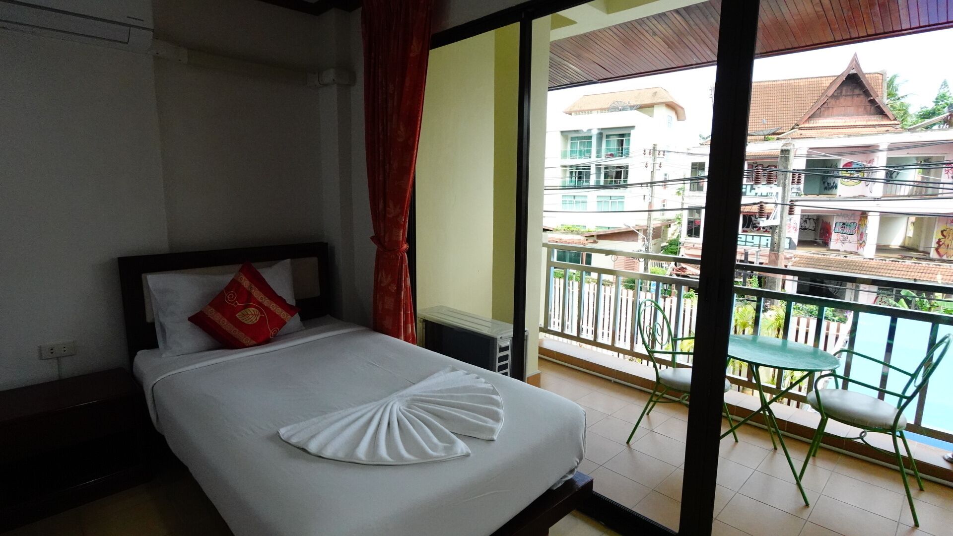A hotel room with a bed, a red pillow, a white towel, and a balcony with chairs and a view of buildings.