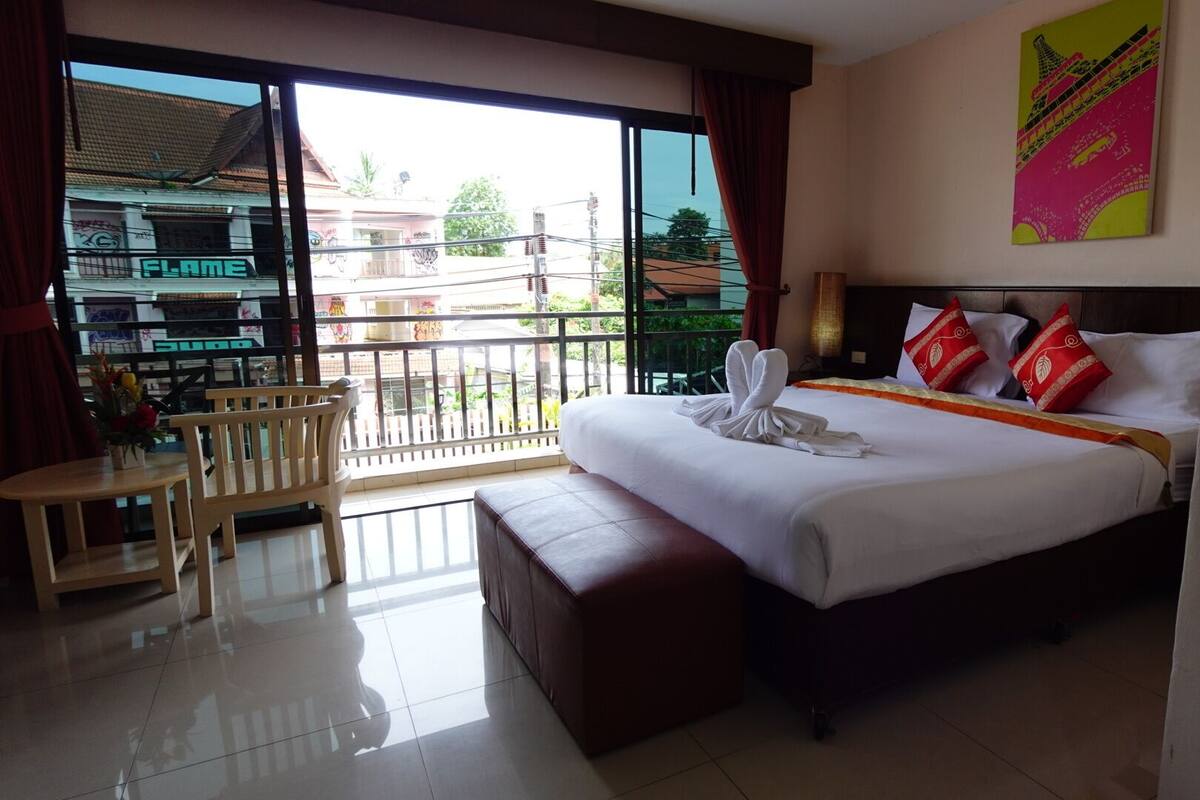 A hotel room with a bed, a small table, a bench, and a balcony with a view of buildings.