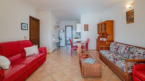 Un salon avec un canapé rouge, un canapé à motifs, une table à manger avec des chaises, et une cuisine en arrière-plan.