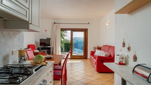 Une cuisine moderne avec une cuisinière à gaz, une table à manger et des chaises rouges. On y trouve un canapé rouge et une petite table avec un grille-pain. Une fenêtre avec un rideau est visible, et une bouteille de Coca-Cola est posée sur une étagère.