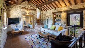 A stone-walled living room with a flat-screen TV, a wooden bench, a patterned rug, a leather armchair, a white sofa with colorful cushions, and a bookshelf. - Grano - Casa Bella Vista (Force)