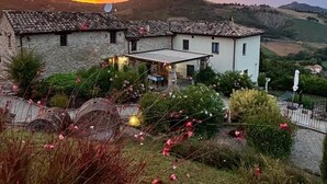 A rustic building with stone walls and a tiled roof, surrounded by a garden with flowering plants and a view of rolling hills in the background. - Grano - Casa Bella Vista (Force)