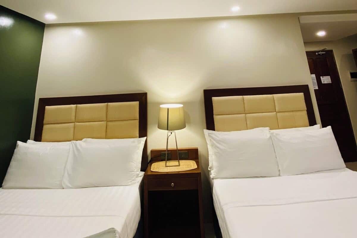 Une chambre d’hôtel avec deux lits, une table de chevet avec une lampe et une porte visible au fond.