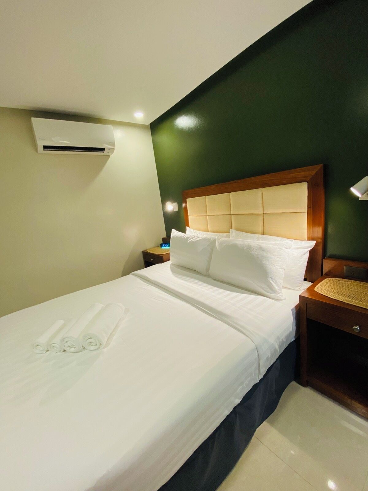 A neatly made bed with white linens and rolled towels, a wooden headboard, a nightstand with a lamp, and an air conditioning unit on the wall.