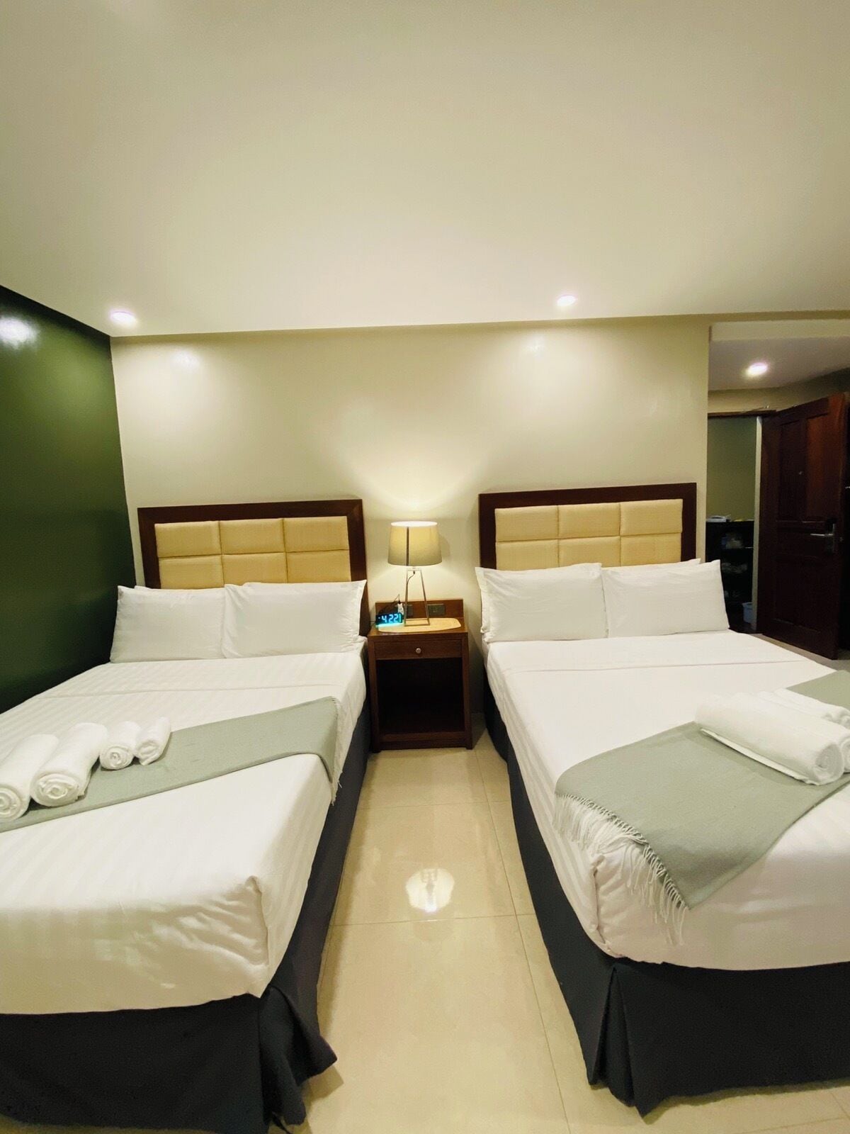 A hotel room with two beds, a nightstand with a lamp, and a digital clock.