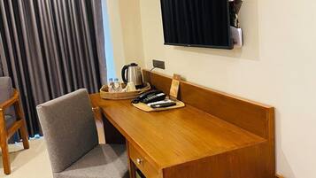 A room with a wooden desk, a grey chair, a television mounted on the wall, and a small table with a kettle and remote controls.