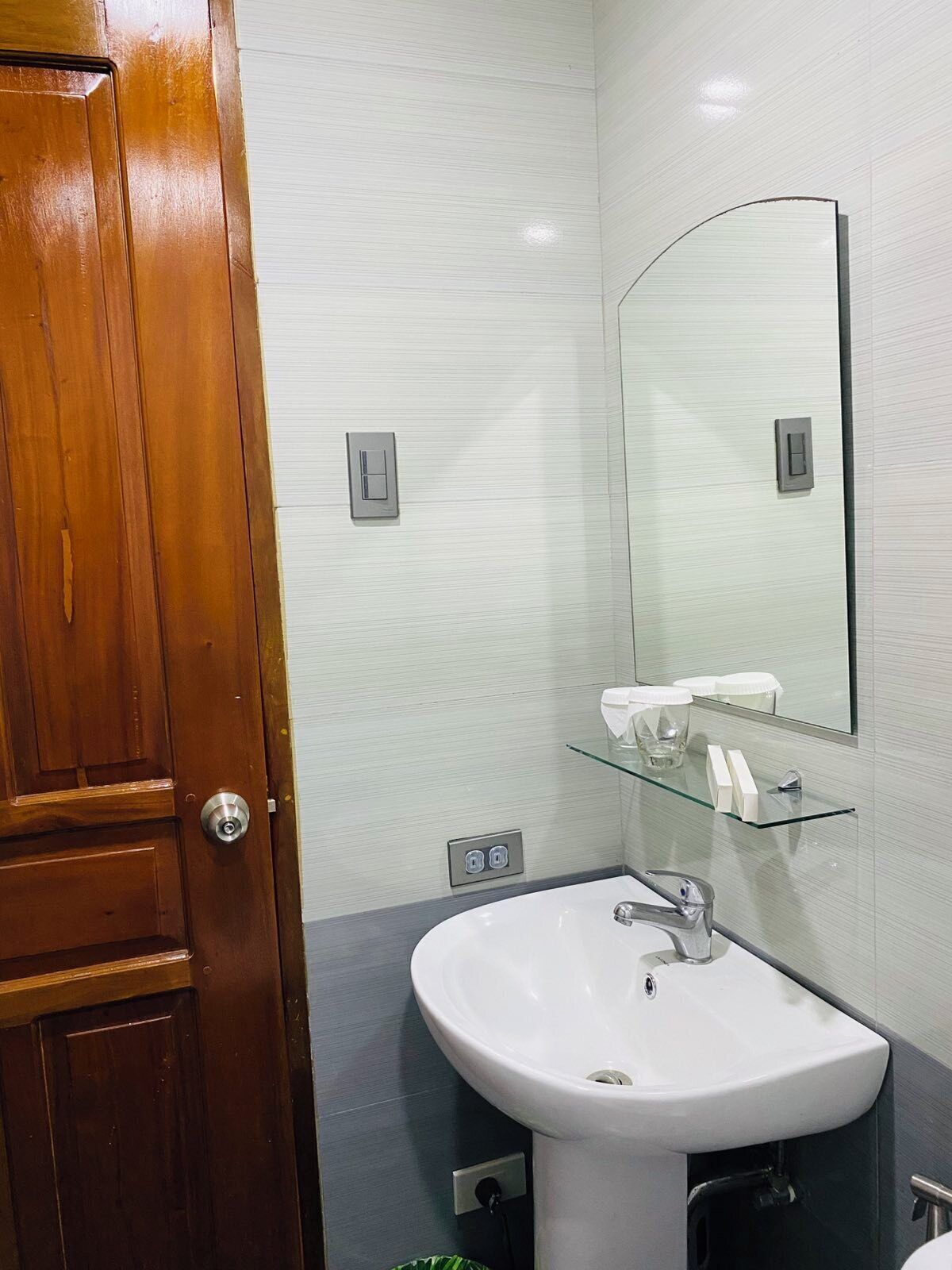 Un baño con puerta de madera, un lavabo blanco, un estante de vidrio con artículos de aseo y un espejo.