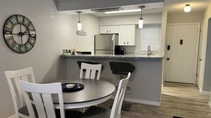 A modern kitchen with white cabinets, a stainless steel refrigerator, and a black countertop. There is a dining area with a round table and white chairs. A wall clock is visible on the left. - Siesta Key Escape: Spacious 2BR /2BA Near Beach + Dining (Siesta Key)