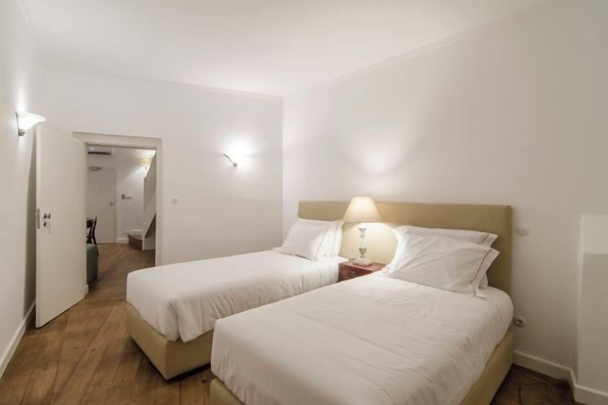 Una habitación de hotel con dos camas individuales, piso de madera, paredes blancas y una mesita de noche con lámpara.