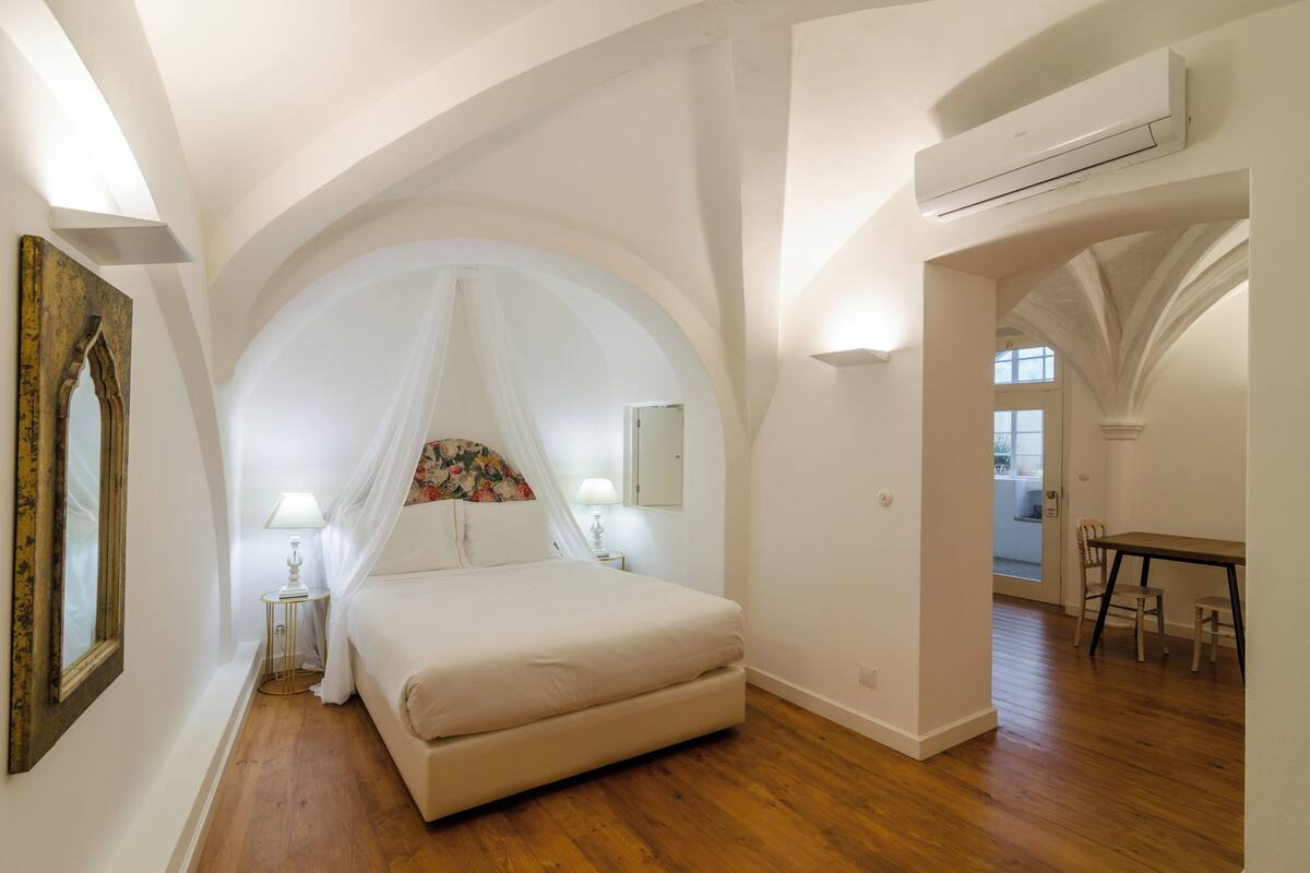 A bedroom with a large bed, a nightstand, a lamp, a mirror, and an arched ceiling.