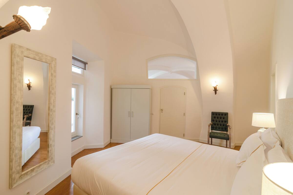 A bedroom with a large bed, a chair, a wardrobe, and a mirror.