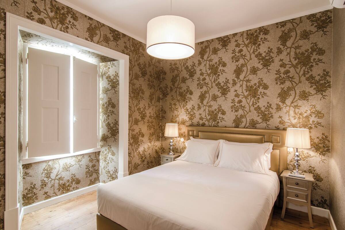 A bedroom with a bed, two bedside tables, a nightstand, a door, and floral wallpaper.
