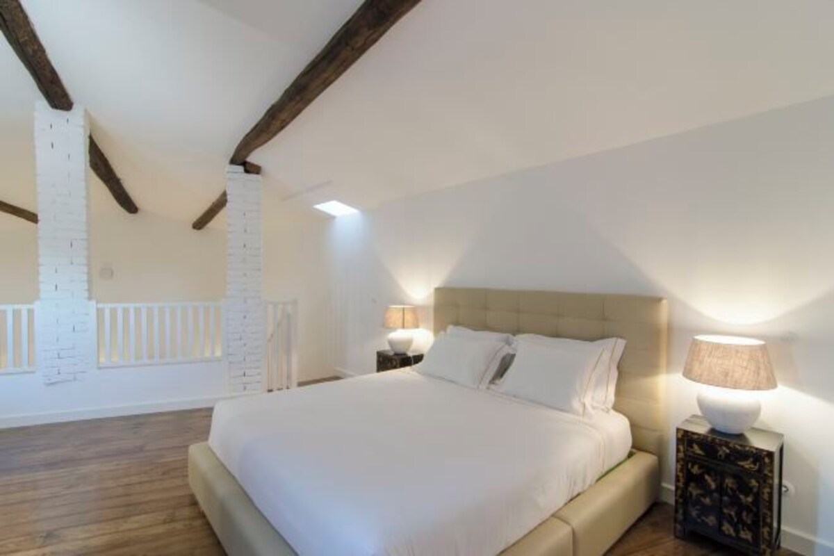 Un dormitorio con una cama grande, vigas de madera, una pared de ladrillo blanco y una mesita de noche con una lámpara.