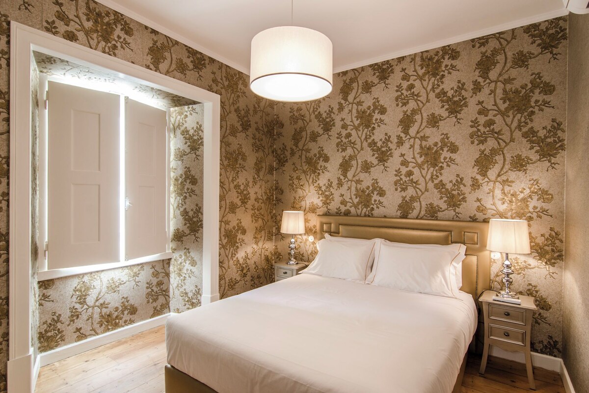 Un dormitorio con una cama, dos mesitas de noche, una mesita auxiliar, una puerta y un papel tapiz con motivos florales.