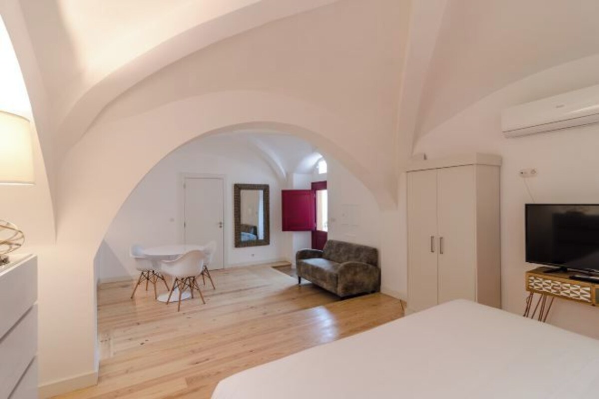 Una habitación amplia con piso de madera, una cama blanca, un sofá, una mesa de comedor con sillas y un televisor sobre un mueble.