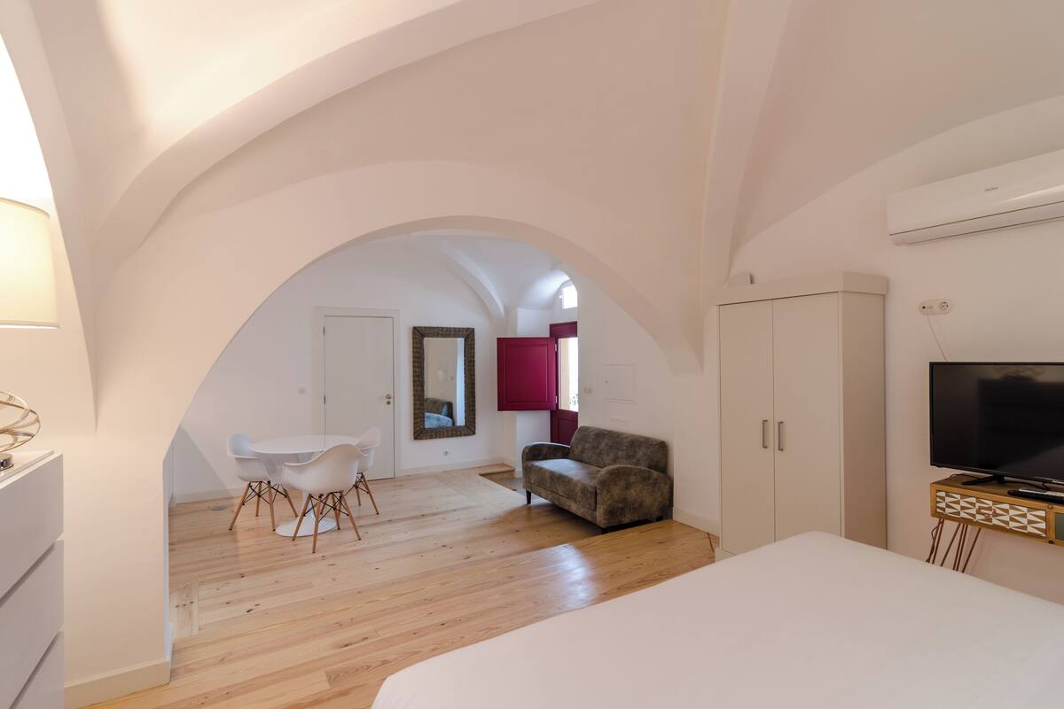 A spacious room with a wooden floor, a white bed, a sofa, a TV on a stand, and a dining area with a table and chairs.