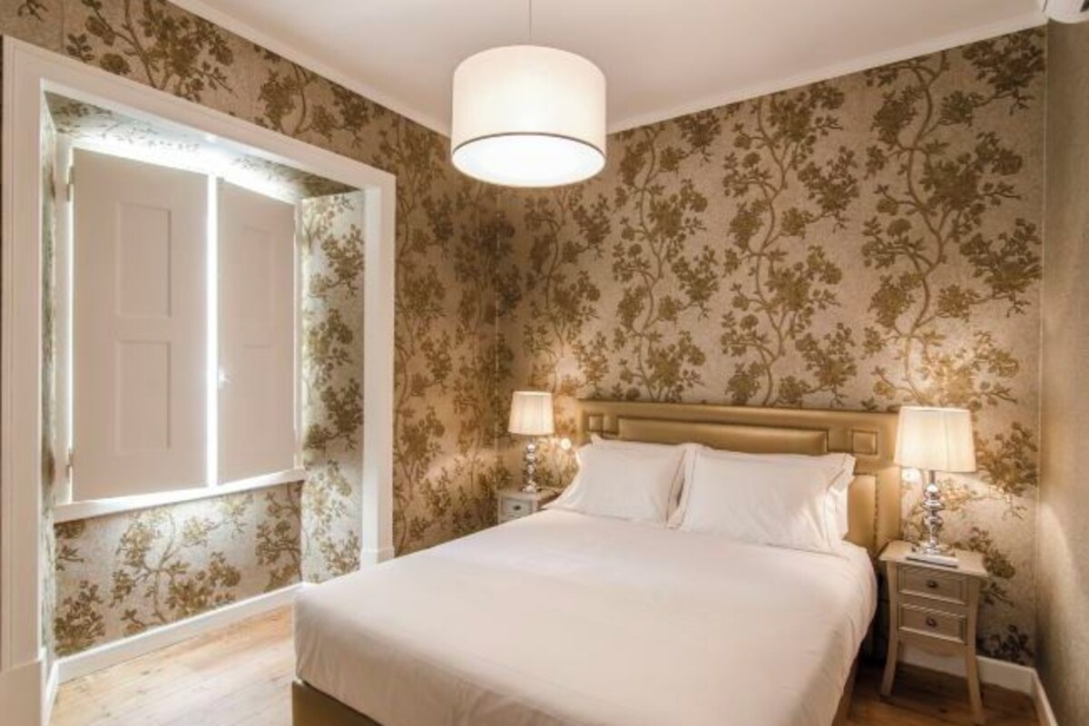 Un dormitorio con una cama, dos mesitas de noche con lámparas, un ventanal con vistas y papel tapiz floral.